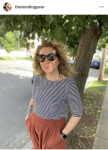 An screenshot of an Instagram post with image of Kate, a white woman smiling at the camera and wearing sunglasses, a black and white striped shirt, and orange linen pants. 